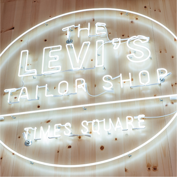 White neon sign reading ‘The Levi’s Tailor Shop — Times Square’ on a wood wall. 