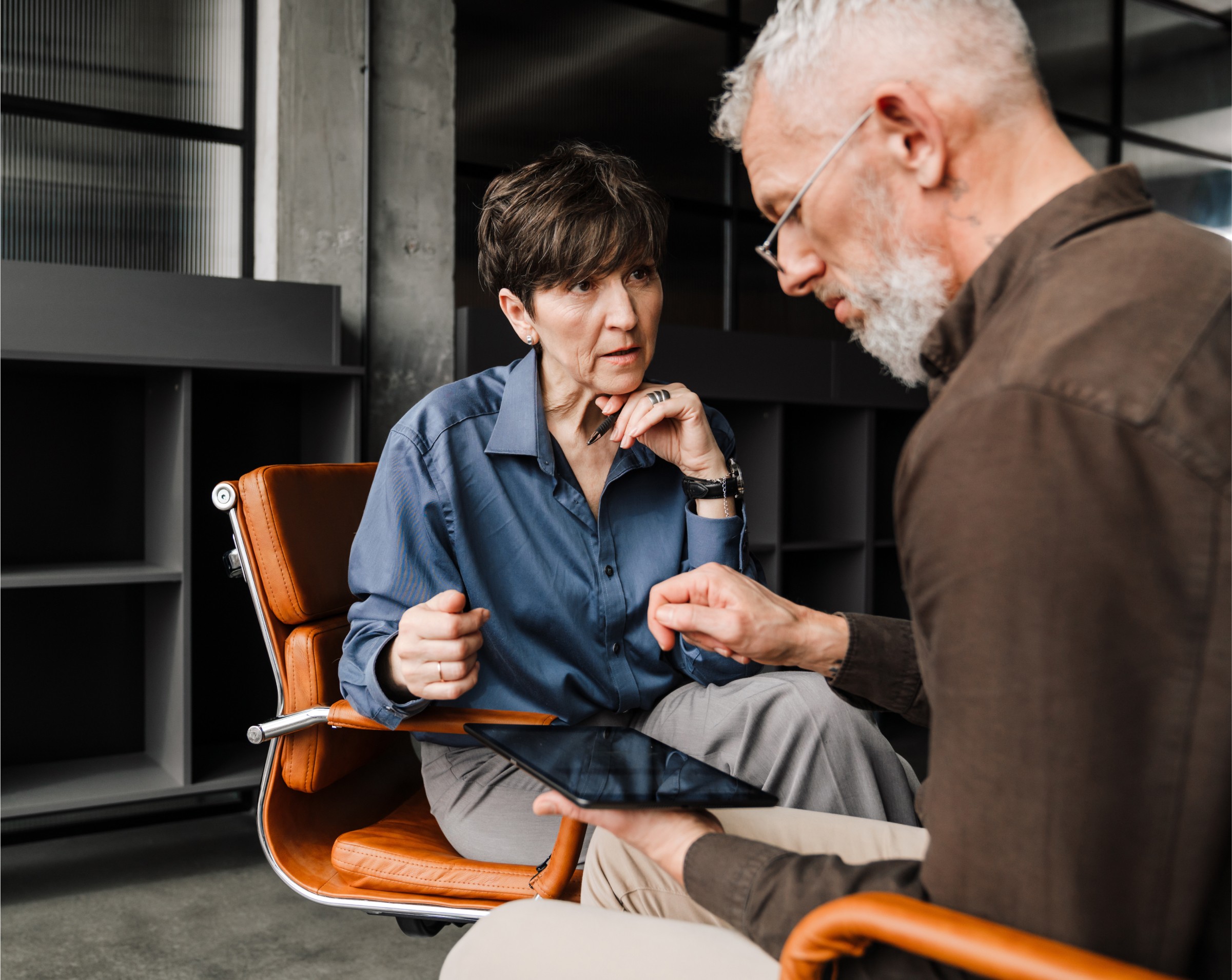 A man and a woman sit in an office, closely reviewing information on a tablet. The woman in a blue shirt listens intently as the man with gray hair explains something