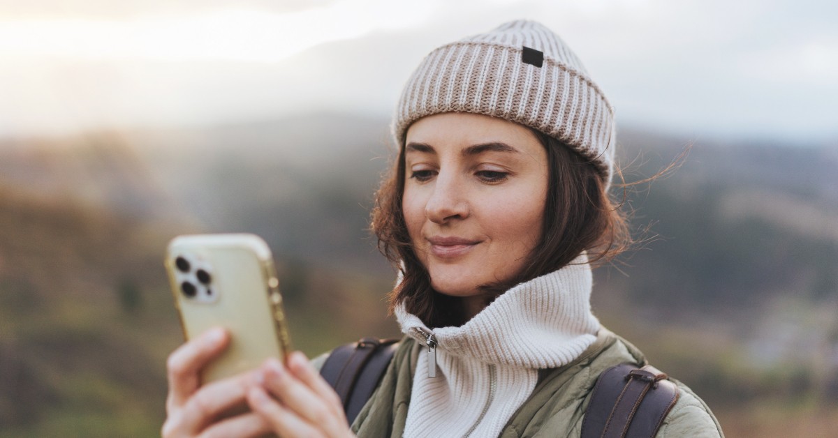 A woman in a knitted beanie and warm jacket, standing outdoors in a scenic area, smiles while looking at her smartphone