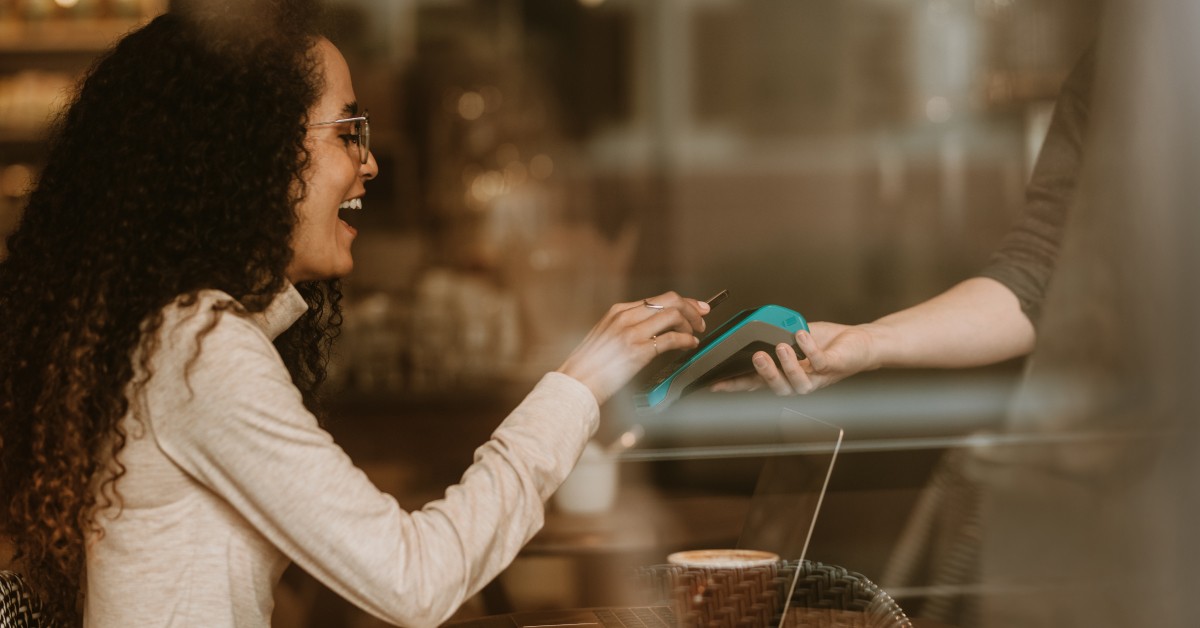A smiling woman with curly hair holding her phone against a card reader to pay at a cafe, while holding a cup of coffee, with a laptop and notebook on the table