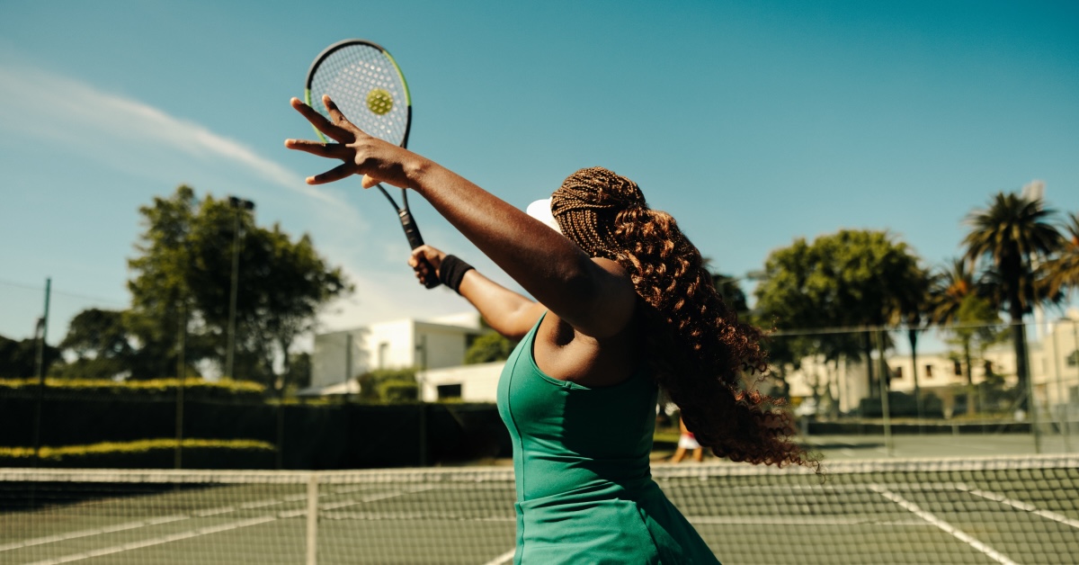 A woman with braided hair is playing tennis on an outdoor court. She is in mid-motion, preparing to serve the ball with her racket. The sun is shining brightly, and the sky is clear, creating a vibrant and energetic atmosphere. The background shows lush greenery and buildings, adding to the picturesque setting. The image captures the athleticism and focus of the tennis player.
