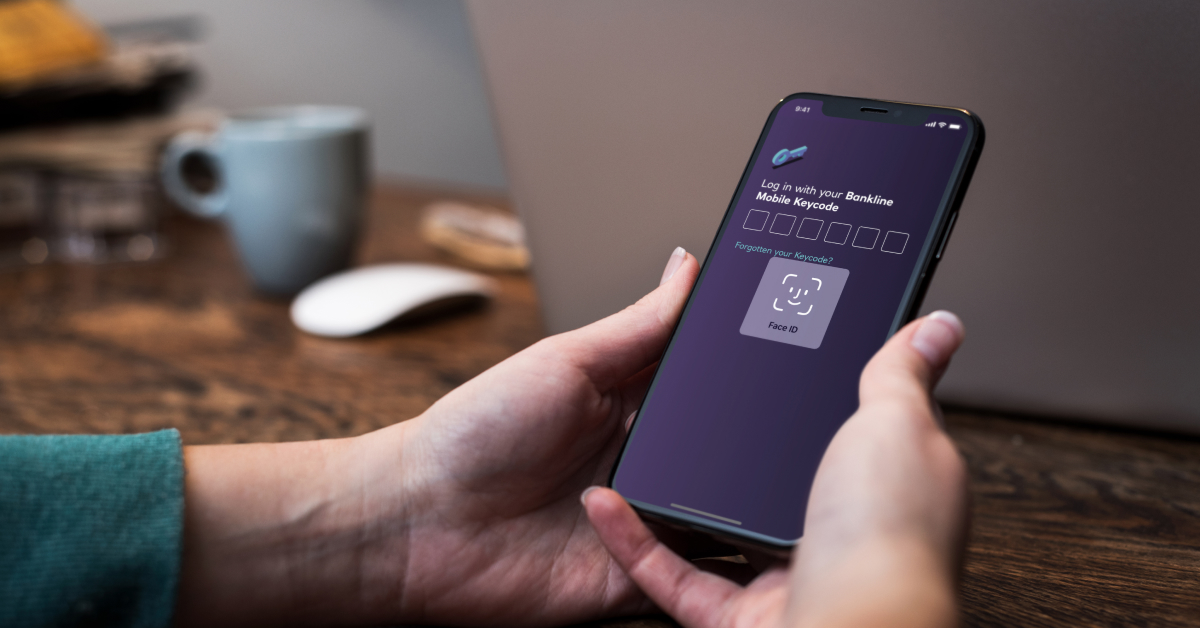 A person holding an iPhone displaying a log in screen. The FaceID functionality of iOS is displayed, together with a heading that reads “Log in with your Bankline Mobile Keycode”, followed by six empty number fields. Below there’s a text link saying “Forgotten your Keycode?”. 